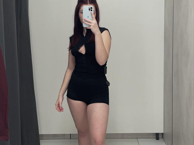 AlexXas with red hair, black outfit, and cutout top, showing her ass and small tits, taking a mirror selfie with a phone in a plain room.