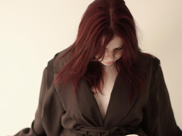 AlexXas with red hair, looking down, wearing a brown, deep V-neck, loose-fitting dress, pale skin, subtle makeup, soft light, slightly wavy hair, focused expression, gentle look.
