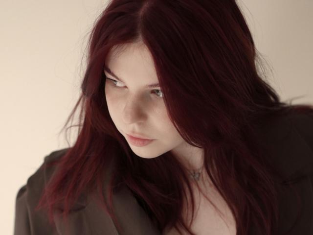 AlexXas has long, dark red hair, pale skin, and a serious expression. She's wearing a black shirt, looking slightly to the side with a subtle, pouty mouth.