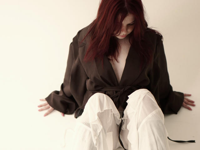 AlexXas with red hair, wearing a brown silk robe and white pants, sitting with hands on the floor, looking down, robe open to reveal a small, bare chest.