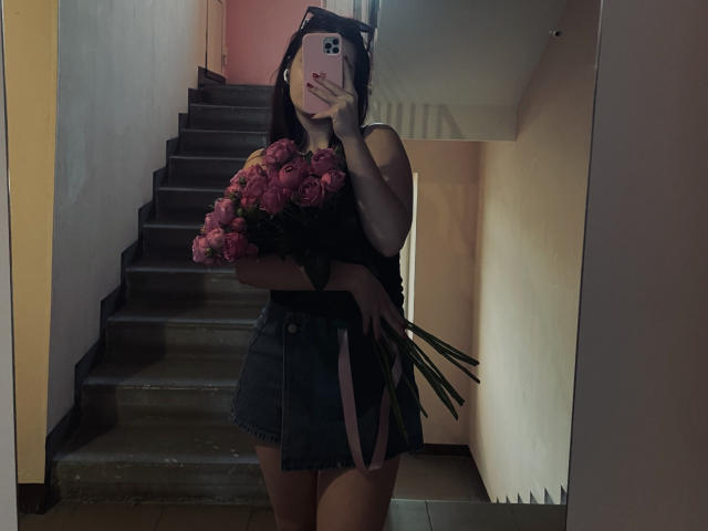 AlexXas with pink roses, holding bouquet in front of her black dress, showing her ass and taut thighs, standing in a dimly lit stairway, taking a mirror selfie.