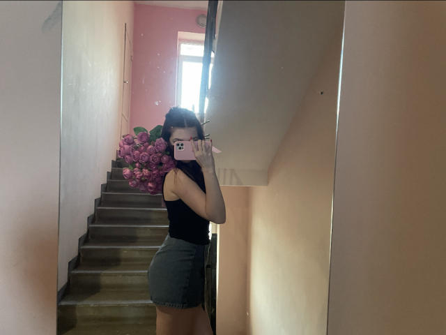 AlexXas has a cute butt in tight green shorts, wearing a black sleeveless top, taking a mirror selfie with a pink phone, standing in a narrow, stair-filled hallway.