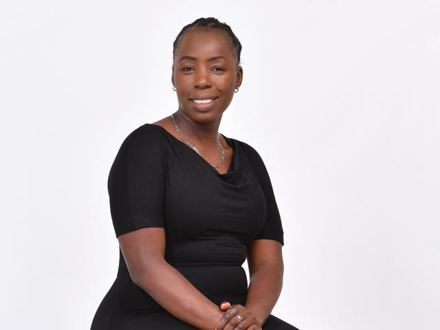 BeutyLicious has dark skin, short braids, and a smile. She's wearing a black dress, silver necklace, and rings. Her tits are medium-sized, and she's sitting.