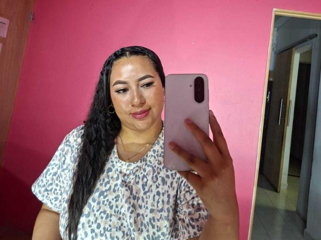 Una ragazza latina con lunghe trecce nere, indossa una camicia a stampa leopardo, tiene un telefono rosa, sorride, mostrando il suo viso luminoso e i suoi glutei piccoli in una stanza con pareti rosa..