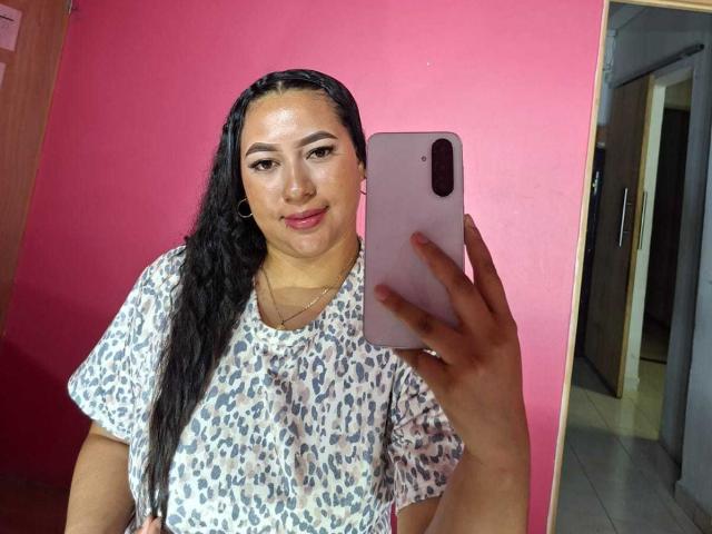 Una ragazza latina con lunghe trecce nere, indossa una camicia a stampa leopardo, tiene in mano un telefono rosa, sorride allo specchio, mostrando il suo viso rotondo e il naso piccolo, sfondo rosa..