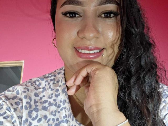 Una mujer latina, con cabello ondulado oscuro, ojos marrones y un piercing en el labio, vestida con una camisa de estampado de leopardo, sonriendo y tocándose la barbilla, con un fondo rosa. Sus senos no son visibles..