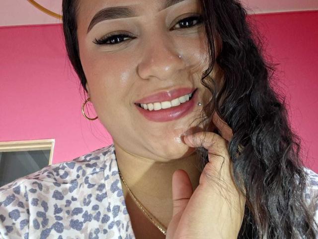 Una mujer latina de cabello negro ondulado, con aros de oro, una camisa de estampado de leopardo, un piercing en la nariz y un piercing en el labio, sonriendo, sosteniéndose la barbilla, mostrando sus dientes y sus labios rosados y brillantes..