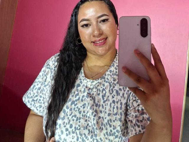 Eine lateinamerikanische Frau mit langen, schwarzen Haaren, die ein leopardenmuster-Shirt trägt, ein rosa Handy hält, lächelt, ihren runden Hintern zeigt und goldene Creolen trägt, in einem Selfie..