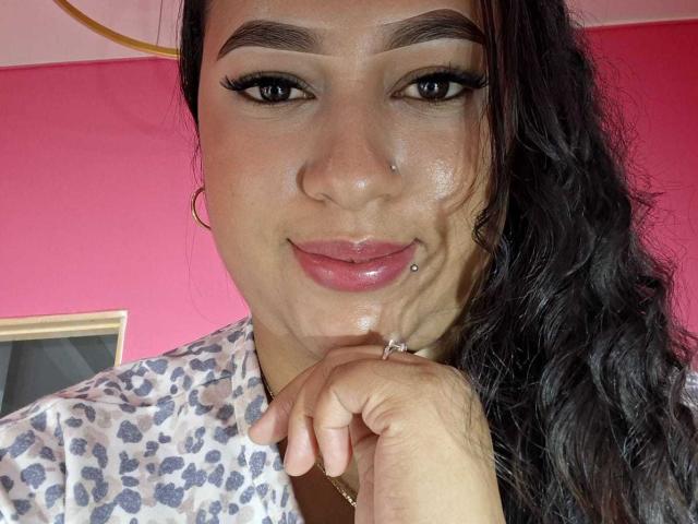 Une femme latina à la peau brillante, aux sourcils foncés, aux longs cils, avec un piercing au nez, un piercing aux lèvres et des cheveux noirs ondulés, portant une chemise à motifs léopard, souriant doucement..