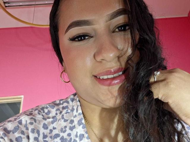 Una donna latina, con capelli scuri e ondulati, sopracciglia folte, rossetto rosa, orecchini a cerchio, una camicia a stampa leopardo, che si tiene i capelli, sorridente, con un anello al dito, carina, attraente, sexy, con la pelle abbronzata..