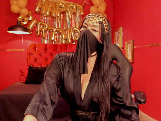 Anissa Ali, vêtue d'une robe en satin noir avec un décolleté plongeant, un masque noir et une pièce de tête dorée, assise sur une chaise noire, dans une pièce rouge, avec des ballons dorés et un lit rouge capitonné derrière elle..