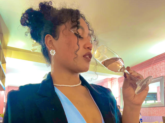 Sophie Sonya, avec ses cheveux bouclés, portant une chemise bleue, un blazer noir, un collier de perles et des boucles d'oreilles en forme de marguerites, sirote du vin rosé dans un verre, en regardant sur le côté..