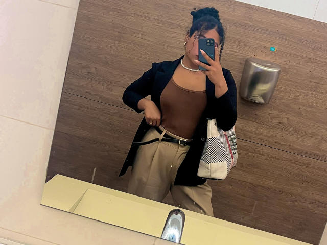 SophieSonyas mit braunen Brüsten, die ein braunes Oberteil, eine schwarze Jacke und eine beige Hose tragen, mit einer Tasche in der Hand, die ein Spiegel-Selfie in einem Badezimmer mit Holzwänden macht..