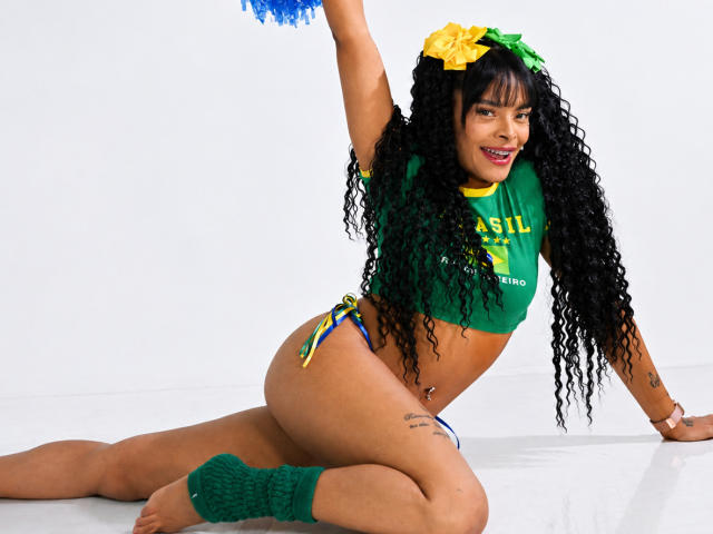EmmaEbony with curly black hair, green top, yellow bow, blue panties, green leg warmers, holding blue pom-poms, smiling, showing her tits and ass, kneeling on white floor.