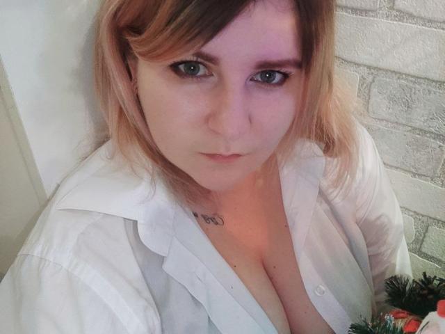MinistraX with pale skin, blue eyes, and blonde hair, wearing a white shirt, showing her tits, looking up at the camera, tattoo above her cleavage, brick wall background.