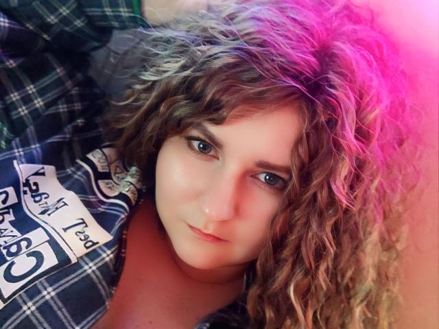 MinistraX has curly brown hair with pink highlights, blue eyes, and a slight smile, wearing a plaid shirt, looking at the camera with a relaxed, confident expression.