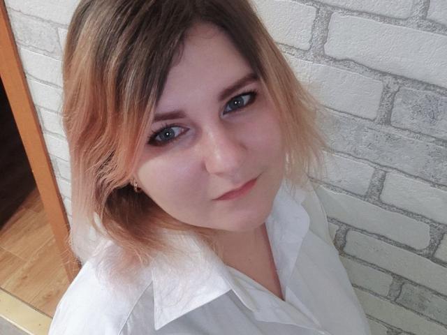 MinistraX has light brown, shoulder-length hair, blue eyes, and a slight smile. She's wearing a white shirt, with a soft, pale complexion and minimal makeup.