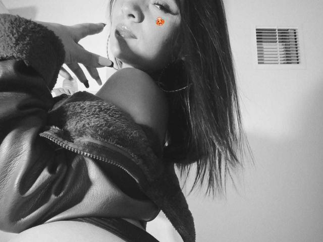 Black-and-white photo of MilaDreams with long dark hair, wearing a furry leather jacket, slightly pulling it down to reveal her bare shoulders and a hint of her ass.