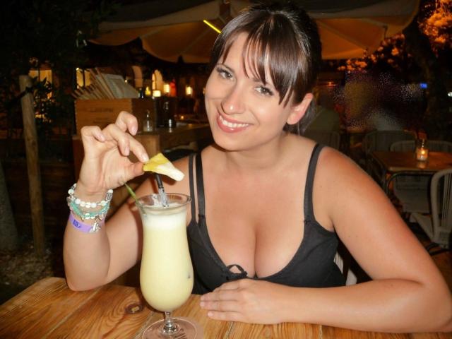 LaLeticia - atractiva, con cabello castaño, sonriendo, sosteniendo una bebida amarilla con una rodaja de limón, vestida con un sostén negro, mostrando sus senos, una mesa de madera y pulseras..