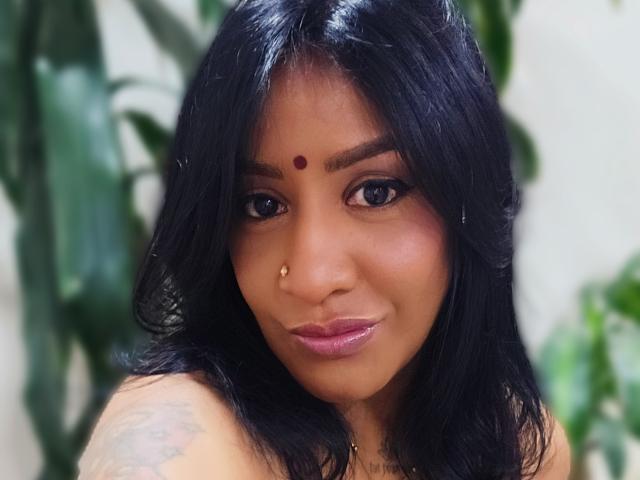 Nahirr hat dunkles, schwarzes Haar, einen roten Bindi auf der Stirn, einen goldenen Nasenring und ein Tattoo an ihrer rechten Schulter. Sie lächelt mit glänzenden, rosafarbenen Lippen..