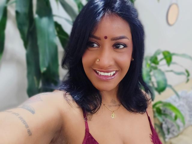 Nahirr, con cabello oscuro, piel morena, un punto rojo en la frente, un piercing en la nariz, sonriendo, vistiendo un sujetador de encaje rojo, un collar, un tatuaje en el brazo, plantas verdes en el fondo..