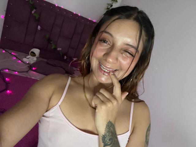 Saarah Jones mit braunen Haaren, die zwinkert, lächelt, mit einem Finger an der Lippe, trägt ein pinkes Tanktop, hat ein Tattoo am rechten Unterarm, ein pinkes Bett, ein violettes Kopfteil, violette Lichterketten, eine weiße Wand..