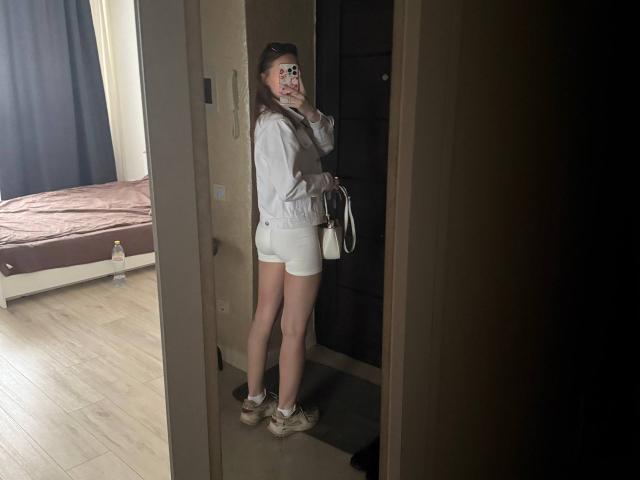 AnyaRuivo prenant un selfie devant un miroir, portant une veste blanche, un short blanc et des baskets, tenant un sac à main, montrant ses fesses, avec une bouteille d'eau sur le lit en arrière-plan..