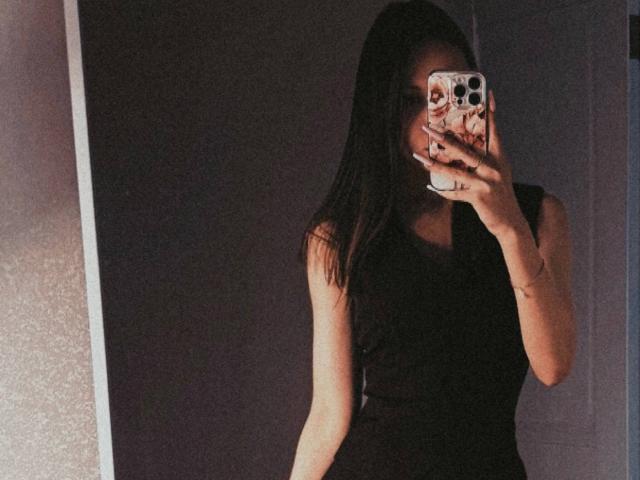 AnyaRuivo with long black hair, wearing a black sleeveless dress, taking a mirror selfie, her phone with a pink patterned case covering her face, showing her tits.