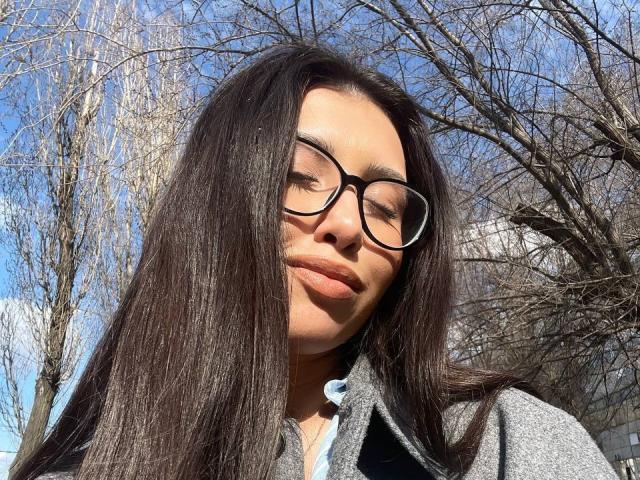 NecoleArciniega has long, straight black hair, wearing glasses and a gray coat, with her eyes closed and a slight smile, standing outdoors with leafless trees in the background.