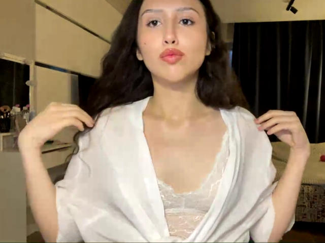 NecoleArciniega with pouty lips, brown wavy hair, white lace bra, and white silky robe, holding robe's edges, showing small tits, in dimly lit room.