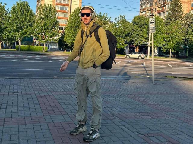 GalaxyGreg, con una sudadera beige, gafas de sol negras, una gorra blanca, pantalones cargo color arena y zapatillas grises, con una mochila negra, de pie en una acera pavimentada con ladrillos..
