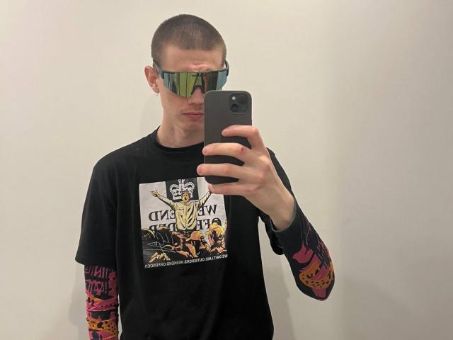 GalaxyGreg, com a cabeça raspada, usando óculos de sol espelhados, camiseta preta, mangas coloridas nos braços, tirando uma selfie no espelho com um smartphone preto..