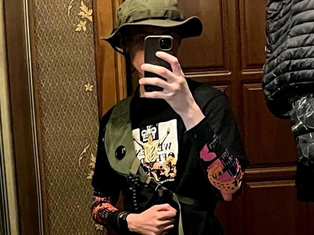 GalaxyGreg mit einem grünen Hut, einem schwarzen Hemd und bunten Ärmeln, der ein Selfie vor einem Spiegel macht, ein Telefon hält und dabei blasse Haut und einen Teil seiner Brust zeigt. Ein skurriles, punkiges Aussehen..