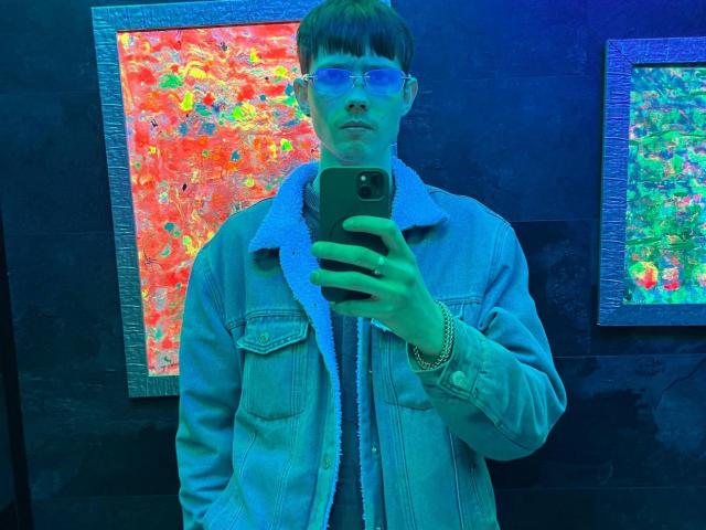 GalaxyGreg mit Brille, Spiegel mit blauen Gläsern, macht ein Selfie in einem neongeladenen Raum. Er trägt eine Jeansjacke, hält ein Telefon, hat ein blaues Armband und kurze, braune Haare..