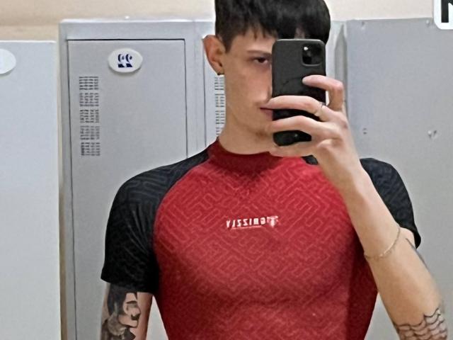 GalaxyGreg with short black hair, wearing a red and black raglan shirt, holding a phone in a mirror selfie, showing tattoos on both arms.