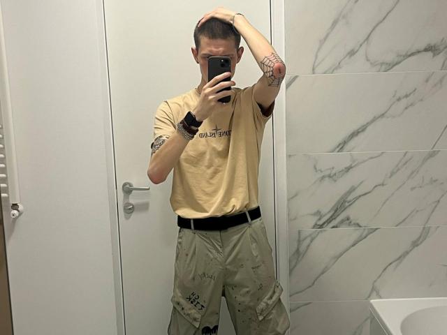 GalaxyGreg, con capelli corti, indossa una maglietta beige, pantaloni cargo verdi, una cintura nera, ha tatuaggi sulle braccia, tiene il telefono per un selfie allo specchio, c'è una porta bianca, piastrelle di marmo, braccialetti, sta guardando il telefono..