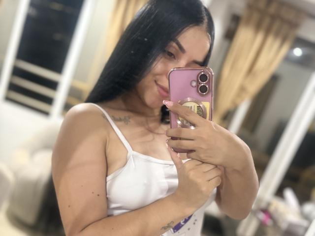 HikariChloe mit langen schwarzen Haaren, weißem Tanktop, die ein rosa Handy hält, ein Selfie macht, lächelt, hat ein sichtbares Tattoo am Hals, kleine Brüste, eine rosa Handyhülle, leicht unscharfes Spiegelbild..