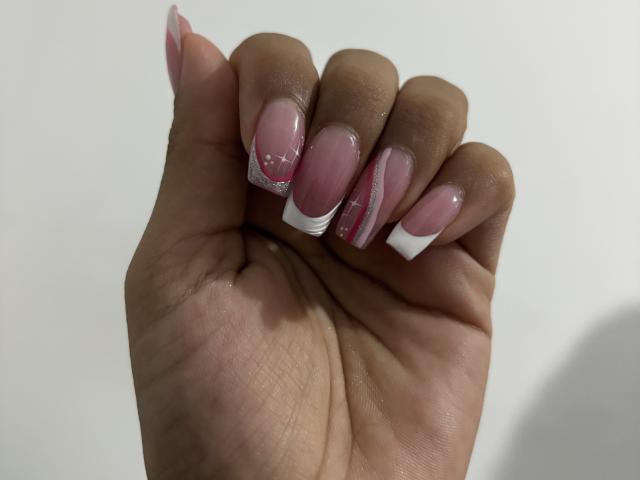 A mão de HikariChloe, com unhas acrílicas rosa e brancas, com pontas transparentes e detalhes brilhantes, está levantada contra um fundo simples. Sua pele é marrom escura e lisa..