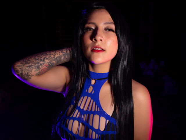 DannyRouds, con lunghi capelli neri, un top di rete blu, un braccio tatuato e rossetto rosso, guarda seducentemente la fotocamera, con i suoi seni parzialmente visibili attraverso la rete. Sfondo scuro..