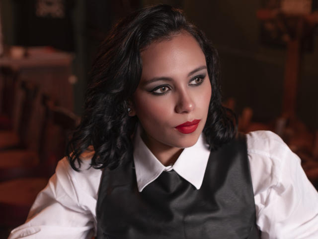 LilithDeveraux has dark wavy hair, red lipstick, and a white shirt with a black leather vest. She's looking sideways with a serious expression. Boobs are small, tits are visible.