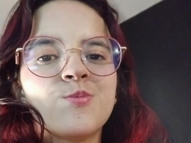 LaiaJonnes with round glasses, red-tinted wavy hair, and pink pouty lips, showing a slight smile. Her nose is centered in the frame.