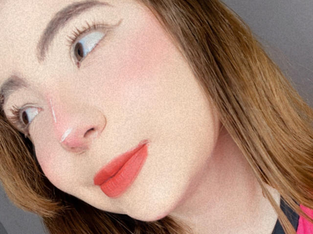 El rostro de Lizy Portman, ligeramente inclinado, con un rubor rosado en las mejillas, lápiz labial rojo y cabello castaño, mirando hacia un lado, mostrando sus seductoras pestañas y su piel suave..