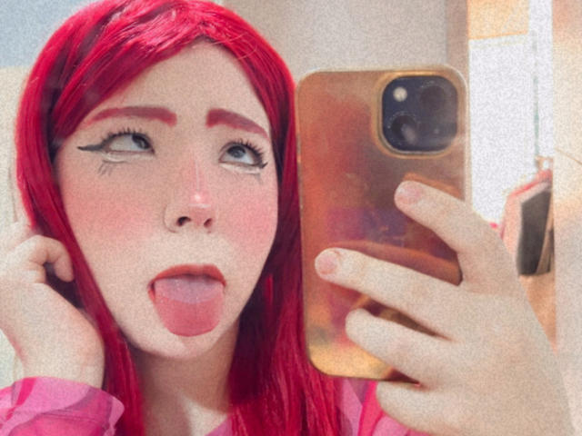 Lizy Portman, avec des cheveux roux éclatants, la langue tirée, tenant un téléphone doré et regardant vers le haut. Elle a des sourcils audacieux, du rouge à lèvres rouge et du crayon noir pour les yeux..