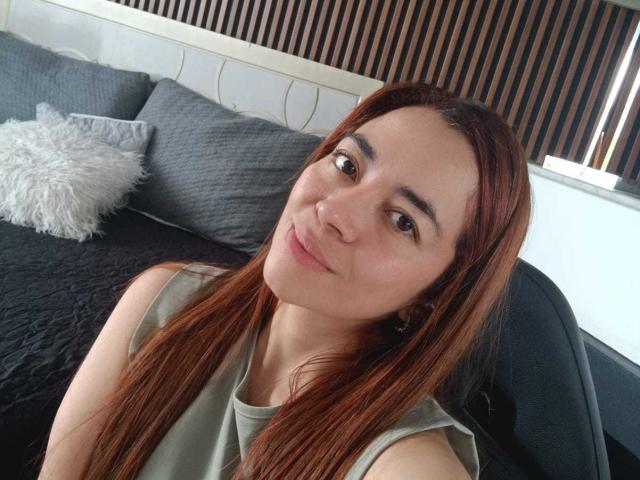 MadisonKoper with long brown hair, light skin, and brown eyes, smiling slightly, wearing a sleeveless gray top, sitting on a black chair, with a gray quilted bed behind.