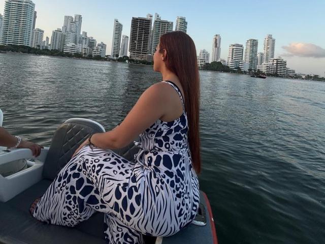 MadisonKoper, with long brown hair, sits on a boat wearing a blue and white leopard-print dress, showing her ass and back, near a city skyline. Water in the background.