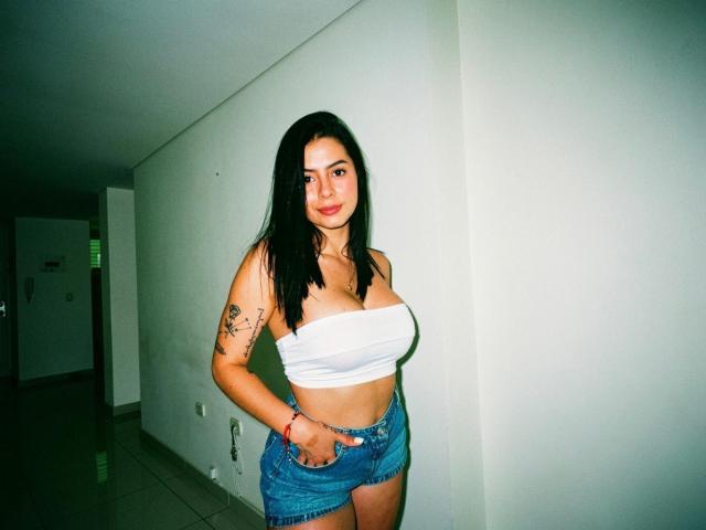 IsabellaJames with black hair, white strapless top, showing big tits, denim shorts, tattoo on right arm, standing in hallway, hands in pockets, smiling, photo with greenish tint.