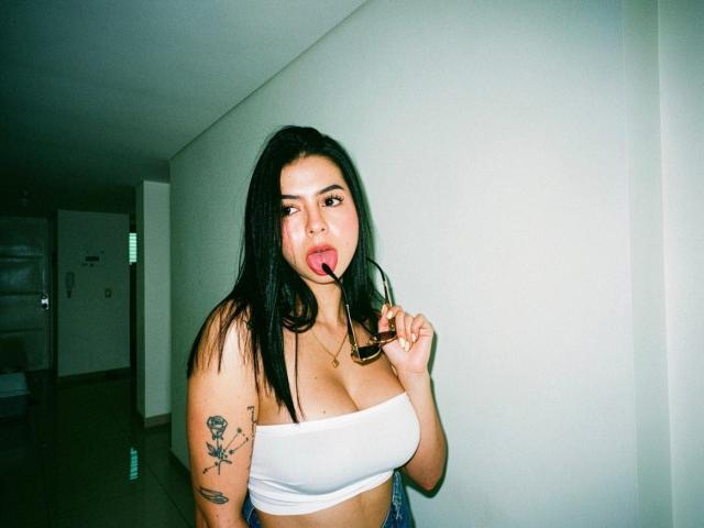 IsabellaJames with black hair, white tube top, licking a lollipop, showing her tits, tattoo on right arm, standing in a hallway, holding glasses with her left hand.