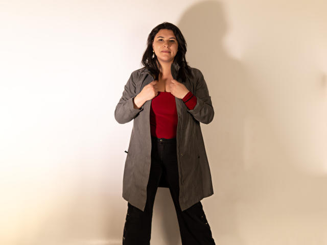 ZoeyGrey with dark hair, red top, gray coat, black pants, standing, hands on coat, shadow on white wall, serious expression, medium build, slightly curvy, small tits.