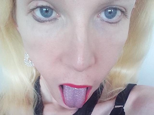 NatachaBradley has blue eyes, blonde hair, and is sticking her purple-tongued face forward with red lipstick, wearing black lace and diamond earrings.