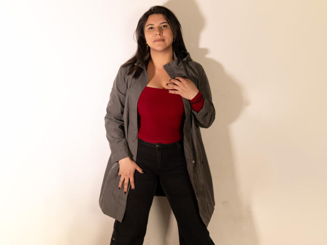 ZoeyGrey stands with a grey coat, red top, and black pants, hand on coat, other hand in pocket, looking confident with a slight smile, medium tits, dark hair.